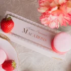 A pink and red box with a clear lid displays six delicate macarons in shades of pink and white. The macarons are perfectly round with a delicate, crisp exterior and a soft, chewy interior. The filling of the macarons is visible through the clear lid, revealing a creamy, pink filling that is flecked with red. A pink ribbon tied around the box adds a touch of elegance and makes it the perfect gift for Valentine's Day. aimee provence